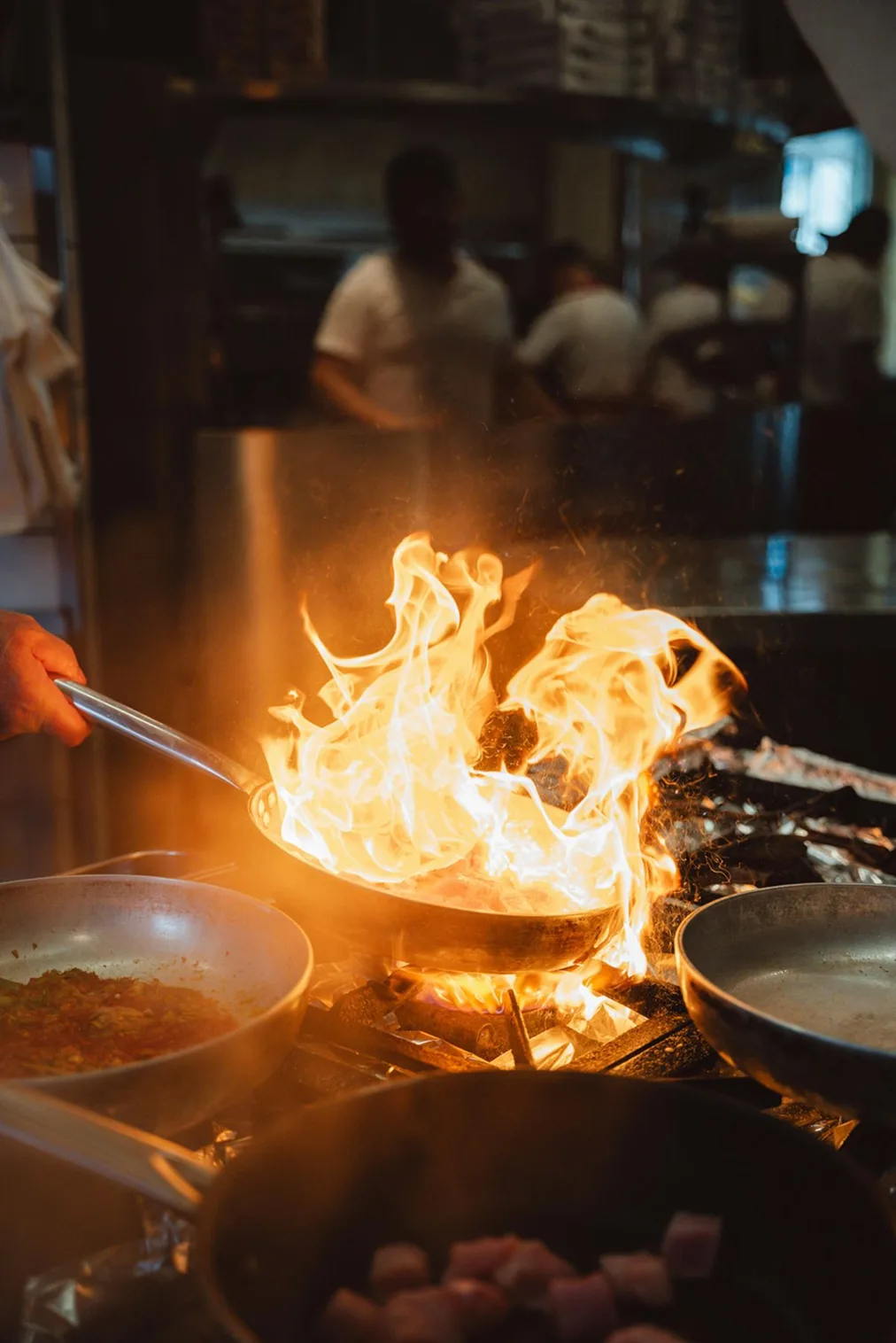 A pan flambé with roaring flames on the stove of a busy restaurant kitchen